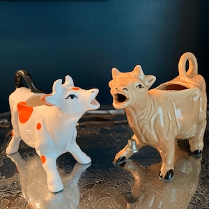 May include: Two ceramic cow-shaped creamers. One is white with orange spots and a black tail handle. The other is solid tan with a small metal bell hanging from its neck. Both cows have open mouths, pointed ears, and tails that form handles.
