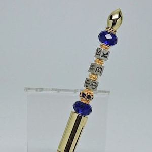 May include: Gold pen with blue and gold beads and a gold top. The beads have the letters "OBK" on them. The pen is on a clear acrylic stand and a black velvet pouch.