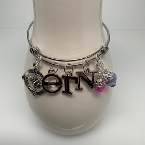 May include: Silver-toned bangle bracelet with charms. Charms include a compass, the letters "OTN", and two beaded orbs in pink and purple. The bracelet is displayed on a white ceramic vase.