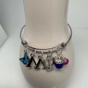 May include: Silver-tone expandable bracelet with multiple charms. Charms include a blue butterfly, the Greek letters Lambda and Psi, and two beaded charms in blue and pink. The bracelet is displayed on a white ceramic vase.