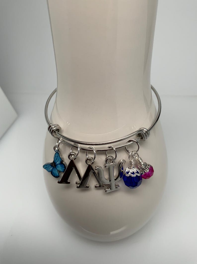 Może przedstawiać: Rozciągliwa bransoletka w kolorze srebrnym z wieloma zawieszkami. Zawieszki obejmują niebieskiego motyla, greckie litery Lambda i Psi oraz dwie zawieszki z koralik&oacute;w w kolorze niebieskim i r&oacute;żowym. Bransoletka jest prezentowana na białym ceramicznym wazonie.