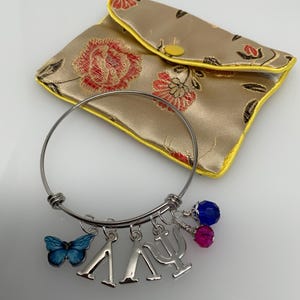 Może przedstawiać: Srebrna bransoletka z zawieszkami, w tym niebieskim motylem, greckimi literami Lambda i Psi oraz zawieszkami z koralik&oacute;w. Złota, kwiatowa torebka z ż&oacute;łtymi wykończeniami znajduje się w tle.