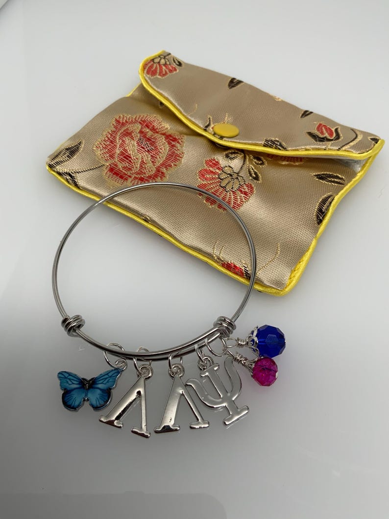 Może przedstawiać: Srebrna bransoletka z zawieszkami, w tym niebieskim motylem, greckimi literami Lambda i Psi oraz zawieszkami z koralik&oacute;w. Złota, kwiatowa torebka z ż&oacute;łtymi wykończeniami znajduje się w tle.