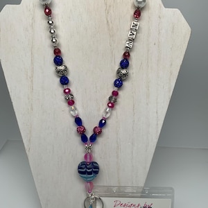 May include: A beaded lanyard with a variety of beads in shades of blue, pink, and silver. The lanyard features a heart-shaped pendant and a butterfly charm. The name "NAVY" is spelled out in silver letters. A clear ID holder is attached.