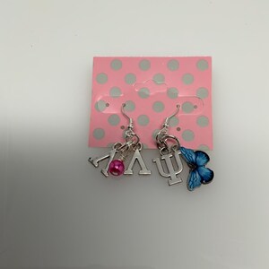 May include: Silver dangle earrings with charms. One earring features the Greek letters Lambda and V, with a pink bead. The other earring has the Greek letter Psi and a blue butterfly charm. The earrings are displayed on a pink card with white polka dots.