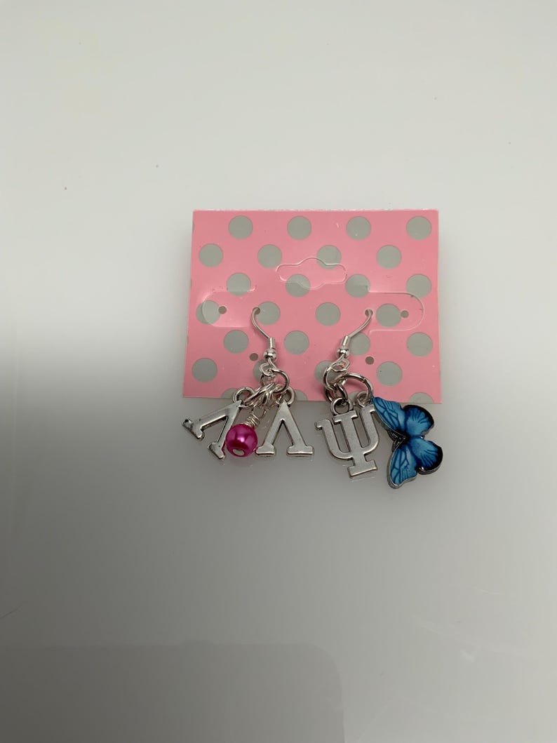 May include: Silver dangle earrings with charms. One earring features the Greek letters Lambda and V, with a pink bead. The other earring has the Greek letter Psi and a blue butterfly charm. The earrings are displayed on a pink card with white polka dots.