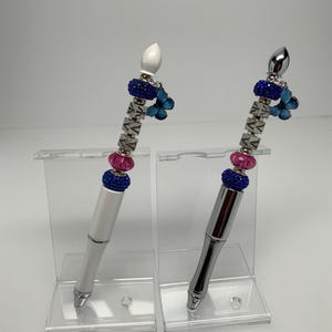 May include: Two decorative pens with silver accents. One pen is white, the other is silver. Both pens have a blue and pink beaded section, a butterfly charm, and a decorative top. The pens are displayed on clear acrylic stands.