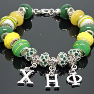 May include: A beaded bracelet with green, yellow, and white beads. The bracelet has silver-toned spacers with green rhinestones and dangling charms with the Greek letters Chi, Heta, and Phi. The bracelet is on a reflective surface.