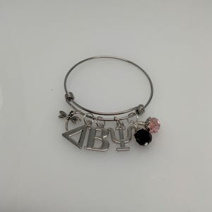 May include: Silver-toned bangle bracelet with charms. Charms include a dragonfly, Greek letters Delta, Beta, and Psi, plus black and pink crystal beads. The bracelet is on a white surface.