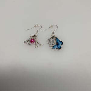 May include: A pair of silver-toned dangle earrings. One earring features the Greek letters Lambda and Mu with a pink bead. The other earring has a blue butterfly charm and the Greek letter Psi.