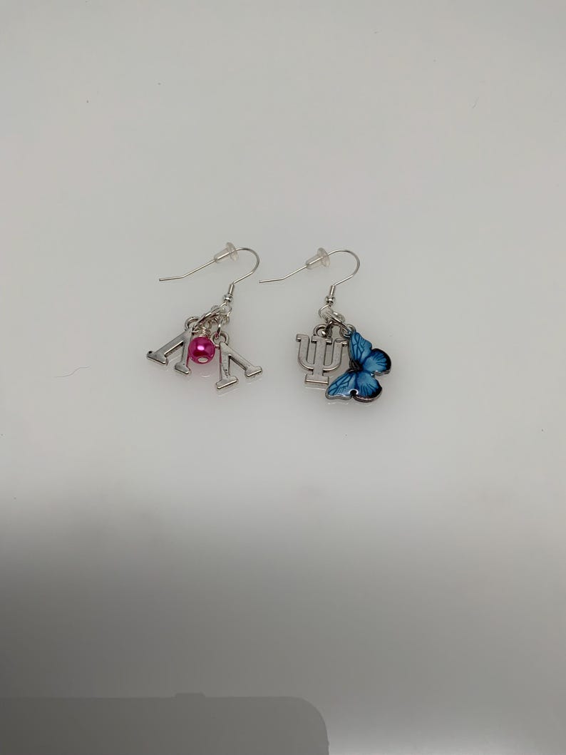 May include: A pair of silver-toned dangle earrings. One earring features the Greek letters Lambda and Mu with a pink bead. The other earring has a blue butterfly charm and the Greek letter Psi.