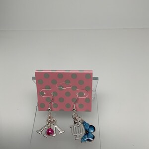 May include: A pair of silver-toned dangle earrings. One earring features a pink bead and a silver charm with the letters "IU". The other earring has a blue butterfly charm and a silver charm with the Greek letter "Psi".