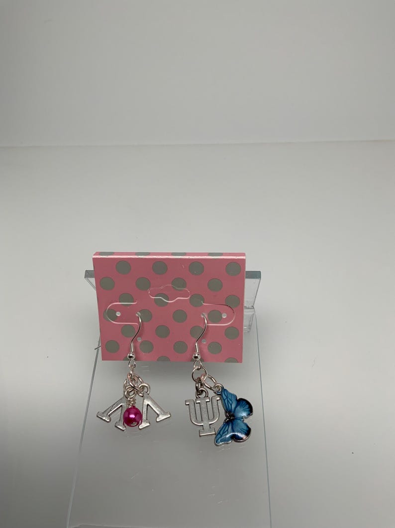 May include: A pair of silver-toned dangle earrings. One earring features a pink bead and a silver charm with the letters "IU". The other earring has a blue butterfly charm and a silver charm with the Greek letter "Psi".