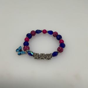 May include: A beaded bracelet featuring a mix of blue, pink, and silver beads. The bracelet includes a blue butterfly charm and silver letter beads spelling out a word. The beads are various shapes and sizes, creating a textured look.
