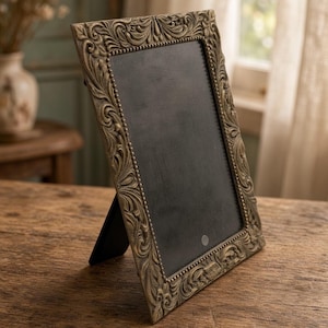 May include: A rectangular tabletop picture frame with an ornate, antique gold-toned finish featuring intricate scrollwork and leaf motifs. The frame has a beaded inner border surrounding a solid black center panel and a black easel-style back support.