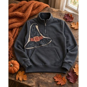 May include: A charcoal gray quarter-zip sweatshirt featuring a large, textured witch hat patch on the chest. The hat is black with a cream-colored outline and a prominent orange buckle. The sweatshirt includes a front kangaroo pocket and ribbed cuffs.
