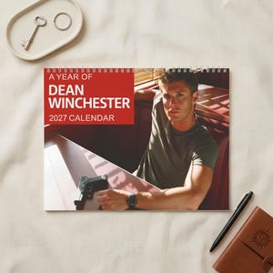 Può includere: Un calendario da parete 2027 intitolato A Year of Dean Winchester con la fotografia di un uomo in maglietta verde seduto in un separé con una pistola sul tavolo. Il calendario presenta una rilegatura a spirale bianca in alto.