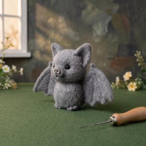 May include: A small, fuzzy, grey needle-felted bat figurine with pointed ears, black bead eyes, and a small snout. The bat has outstretched wings and is positioned on a green surface next to a wooden-handled metal felting needle tool.