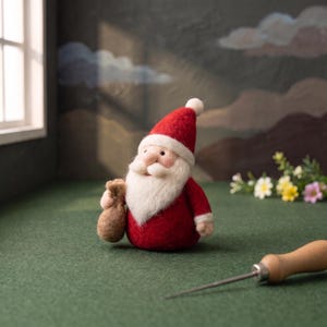 Könnte beinhalten: Eine nadelgefilzte Weihnachtsmann-Figur aus roter und weißer Wolle mit einer spitzen Mütze, einem dichten weißen Bart und einem kleinen braunen Sack in der Hand. Die Figur hat einen abgerundeten Körper und liegt neben einem hölzernen Filzwerkzeug.