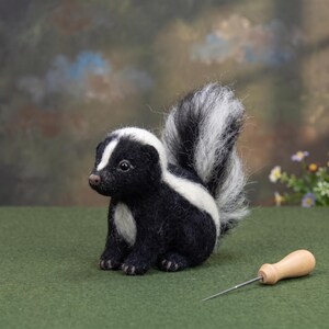May include: A needle-felted skunk figurine with black and white wool fibres, featuring a prominent bushy tail, a white stripe running from the head down the back, and a small brown nose. A wooden-handled felting needle lies beside the figurine.