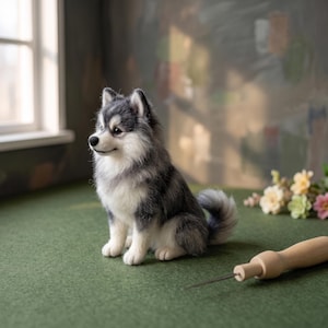 Puede incluir: Una figura de perro en miniatura de lana fieltrada con pelaje gris, negro y blanco, representada en posición sentada con la cola enroscada. Una aguja de fieltrar con mango de madera se encuentra junto a la pequeña y detallada escultura.