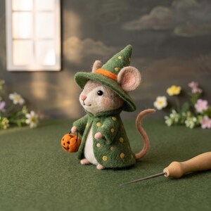 May include: A needle-felted mouse figurine wearing a green pointed hat with an orange band and a matching green cloak featuring yellow polka dots. The mouse holds a small orange jack-o'-lantern bucket and has a long pink tail, with a felting needle nearby.