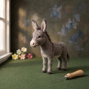 May include: A needle-felted donkey figurine crafted from gray wool fibers, featuring upright ears, a dark mane, and a lighter muzzle. The donkey stands on four legs with dark hooves, positioned near a wooden-handled felting tool on a green surface.