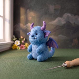 May include: A needle-felted blue monster figurine with purple horns, purple inner ears, and purple wings. The creature has a round body, two small white teeth, and black eyes. A wooden-handled felting tool is positioned in the foreground.