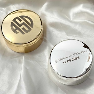 Könnte beinhalten: Zwei runde Schmuckdosen aus Metall mit Deckel. Eine Dose ist goldfarben und zeigt ein kreisförmiges Monogramm mit den Buchstaben ASD. Die andere Dose ist silberfarben und mit dem Text William & Christine sowie dem Datum 11.09.2026 graviert.