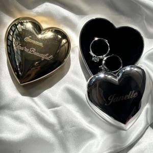 May include: Two heart-shaped jewelry boxes. One is gold-toned with the engraved text 'Summer, You're Beautiful!'. The other is silver-toned, engraved with 'Janelle', and is open to reveal two silver rings with clear stones inside the black interior.