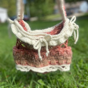 May include: A crochet bag featuring a variegated rust, brown, and cream yarn body with a textured basketweave pattern. The bag includes a cream-colored ruffled trim along the top edge, a matching drawstring tie, and a decorative openwork lace border.