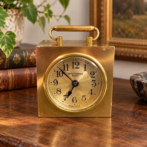 May include: A square brass Seth Thomas 8 Day clock with a handle on top. The round clock face features black Arabic numerals, black hands, and the text Seth Thomas 8 Day and Made in U.S.A. printed in black on the metallic gold dial.