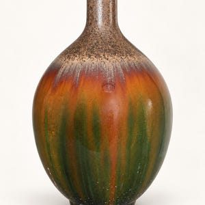 Puede incluir: Un jarrón de cerámica con cuello estrecho y cuerpo redondeado con un esmalte brillante. La superficie muestra un patrón de goteo vertical que transita desde un borde texturizado marrón oscuro hasta vetas de naranja y verde bosque.
