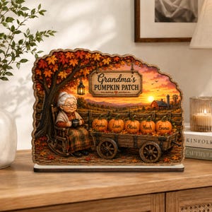 Könnte beinhalten: Ein dekoratives Holzschild mit einer Illustration einer Person mit weißem Haar, die in einem Schaukelstuhl unter einem Baum mit orangefarbenen Herbstblättern sitzt. Ein Holzwagen trägt fünf Kürbisse mit den Namen Sophia, Liam, Olivia, Noah und Emma.