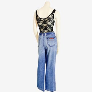 May include: A mannequin displays a black lace bodysuit paired with light-wash denim jeans. The jeans have a flared leg and a small red and white logo on the back pocket. The bodysuit has a scoop back and a scoop neck.
