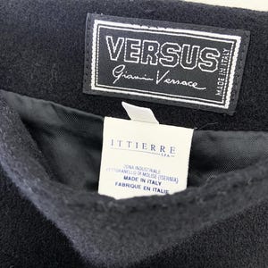 May include: Close-up of a black garment with a Versus Gianni Versace label. The label is white with black text and a stitched border. A second white label with text in Italian and French is visible.