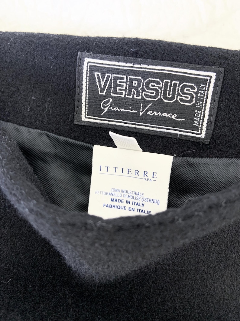 May include: Close-up of a black garment with a Versus Gianni Versace label. The label is white with black text and a stitched border. A second white label with text in Italian and French is visible.