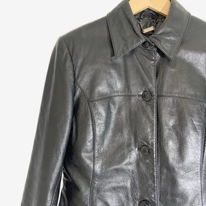 May include: A black leather jacket with a collared neckline and three button closure. The jacket is hanging on a wooden hanger against a white background. The jacket has a classic, tailored design.