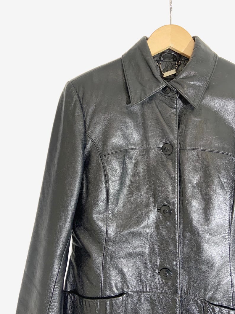 May include: A black leather jacket with a collared neckline and three button closure. The jacket is hanging on a wooden hanger against a white background. The jacket has a classic, tailored design.