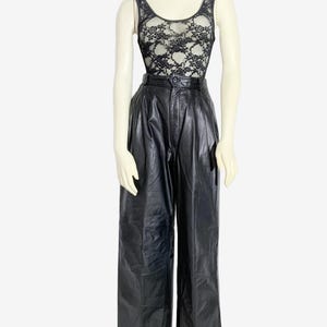 May include: A mannequin showcases a black lace bodysuit and high-waisted, wide-leg black leather trousers. The bodysuit has a floral lace design over a nude lining. The trousers feature a pleated front and a button closure.