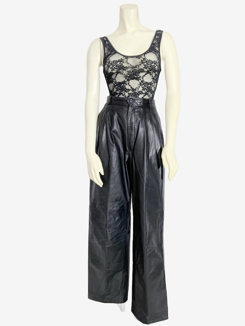 May include: A mannequin showcases a black lace bodysuit and high-waisted, wide-leg black leather trousers. The bodysuit has a floral lace design over a nude lining. The trousers feature a pleated front and a button closure.
