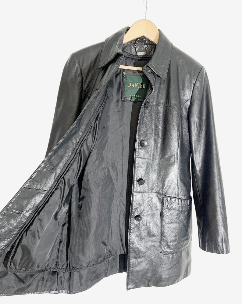 May include: Black leather jacket with a collared neckline, button closure, and two front pockets. The jacket is hanging on a wooden hanger. The interior lining is visible.