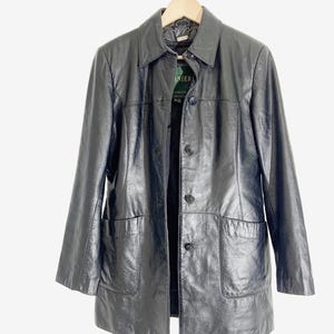 May include: A black leather jacket with a collared neckline, button-up front, and two front pockets. The jacket is hanging on a wooden hanger against a white background. The tag reads "LANIER CANADA".
