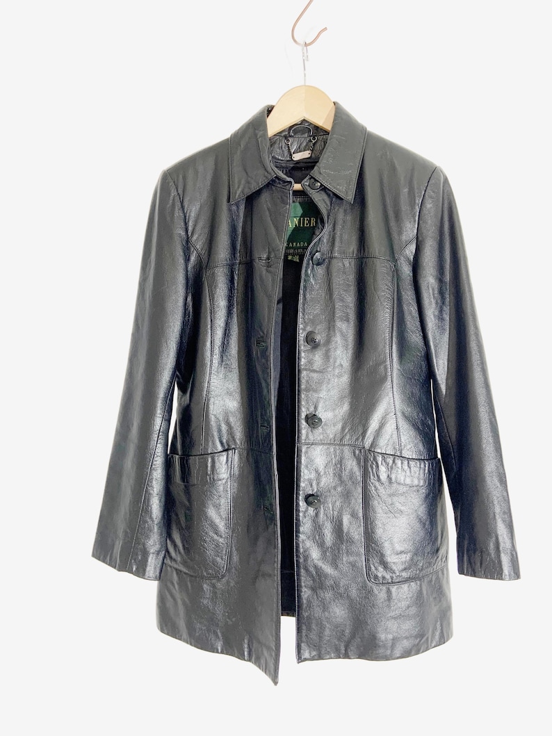 May include: A black leather jacket with a collared neckline, button-up front, and two front pockets. The jacket is hanging on a wooden hanger against a white background. The tag reads "LANIER CANADA".