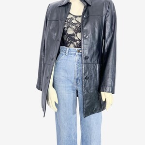 May include: A black leather jacket with a button-up front and a collared neckline. The jacket is worn over a black lace top and light blue jeans. The jacket has a classic, tailored design.