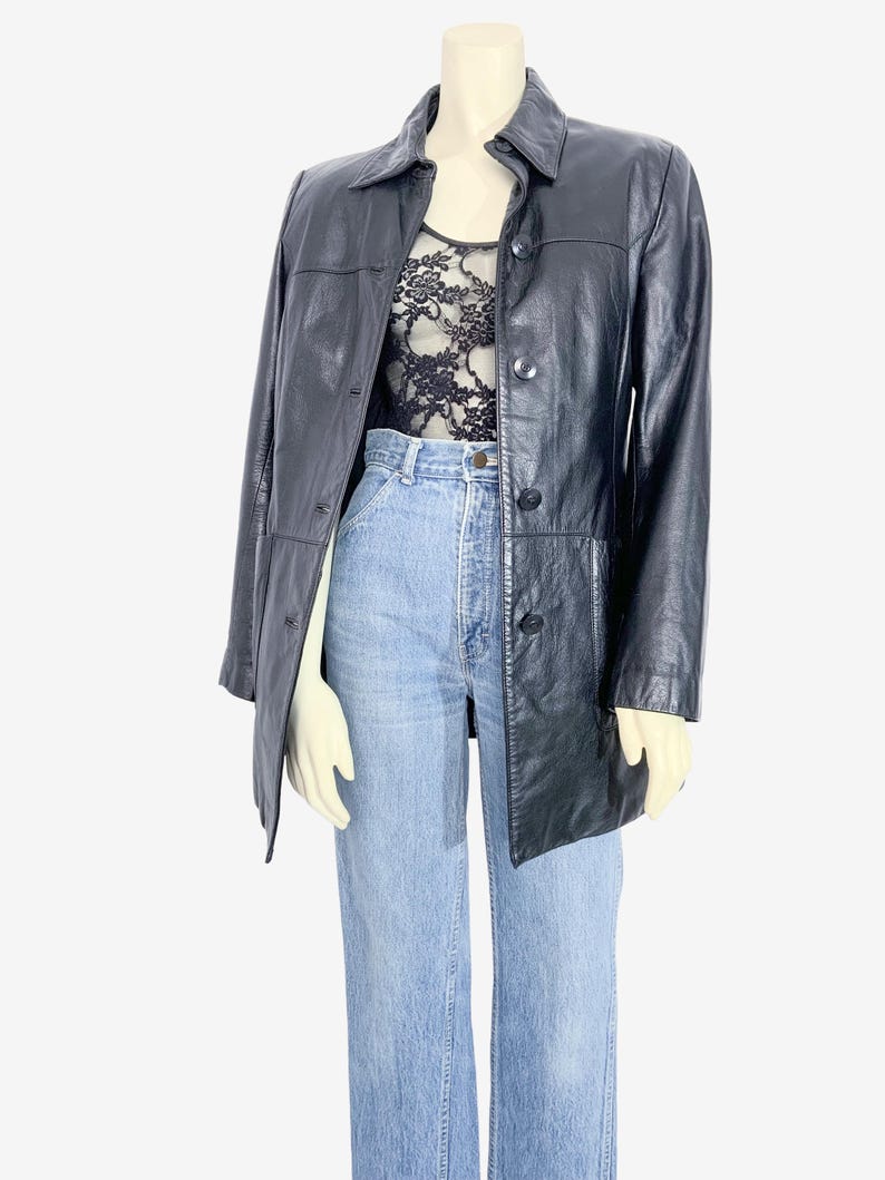 May include: A black leather jacket with a button-up front and a collared neckline. The jacket is worn over a black lace top and light blue jeans. The jacket has a classic, tailored design.
