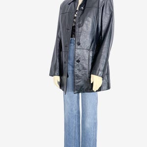 May include: A black leather jacket with a button-up front and a collared neckline. The jacket is paired with light blue denim jeans. The jacket appears to be a vintage or retro style.