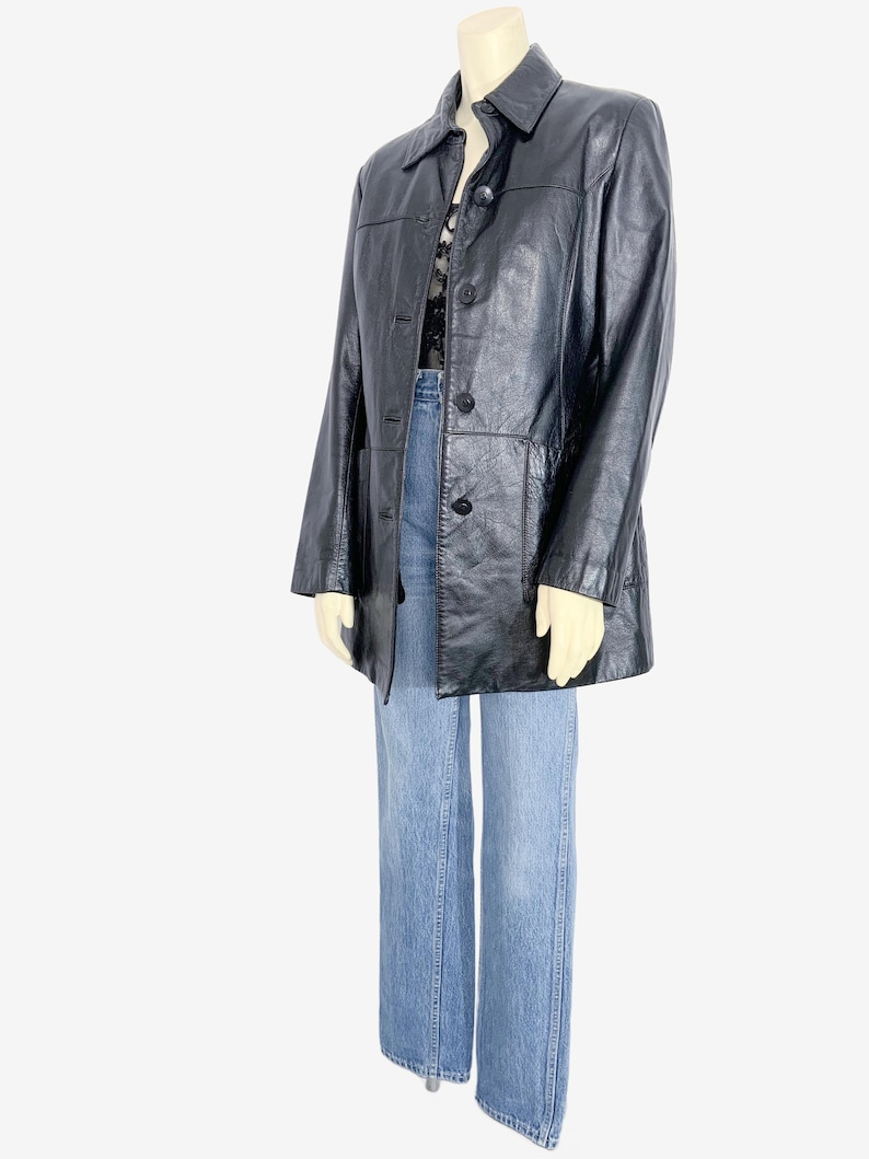 May include: A black leather jacket with a button-up front and a collared neckline. The jacket is paired with light blue denim jeans. The jacket appears to be a vintage or retro style.