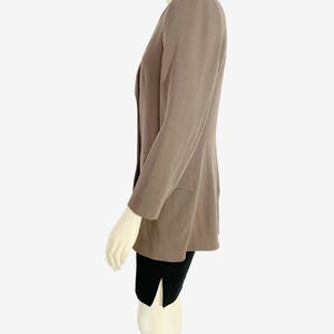 May include: A taupe blazer with a notched lapel and two front pockets. The blazer has long sleeves and is worn over a black garment. The blazer is displayed on a mannequin.
