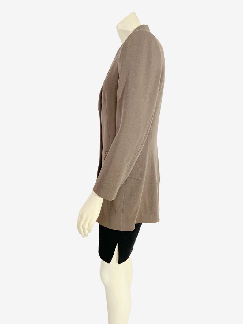 May include: A taupe blazer with a notched lapel and two front pockets. The blazer has long sleeves and is worn over a black garment. The blazer is displayed on a mannequin.